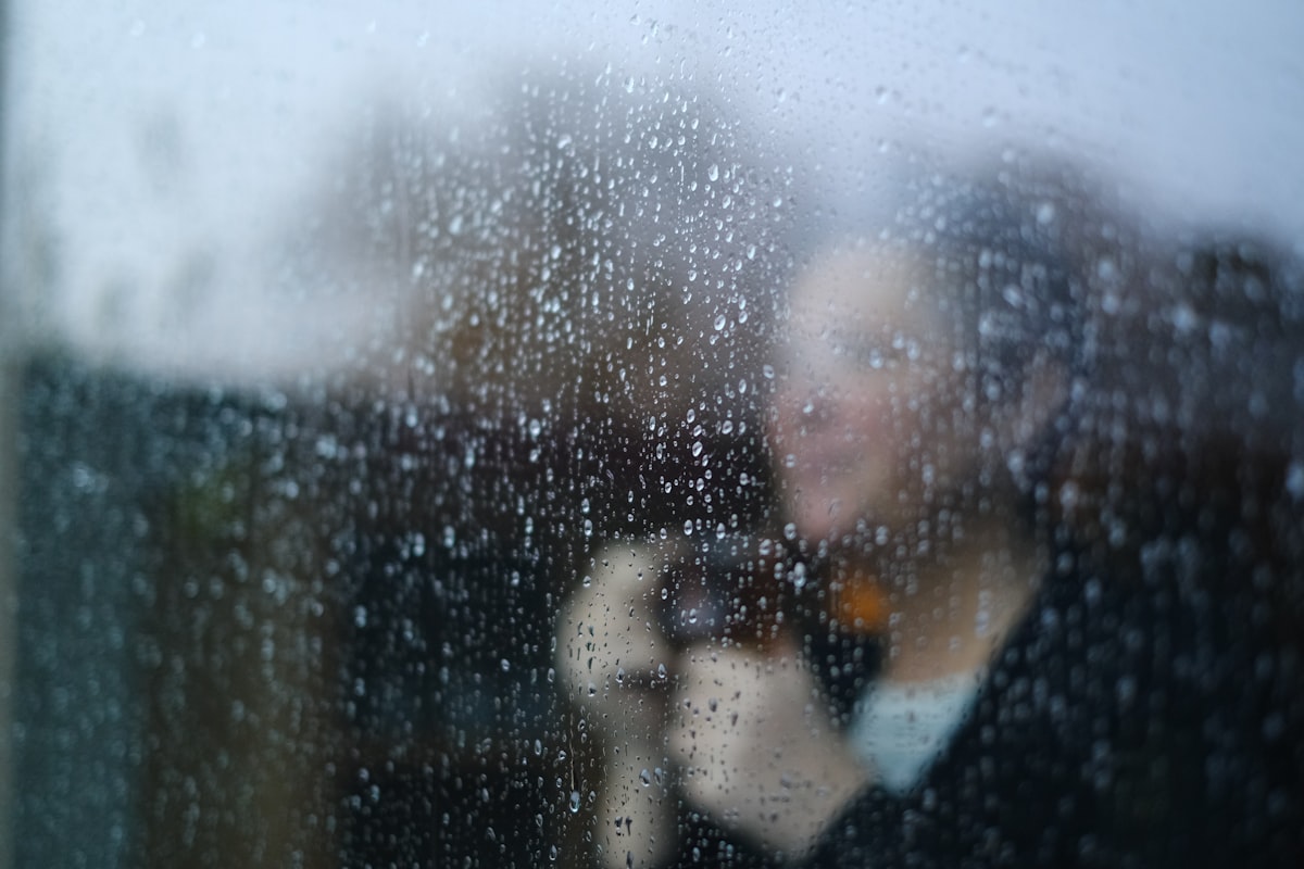 A figure with a warm cup of tea, seen softly through a rain-speckled window
