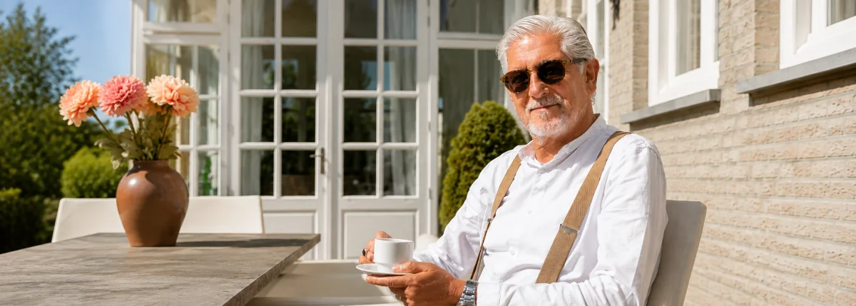 An older man in sunglasses sits outside in the sunshine with a coffee